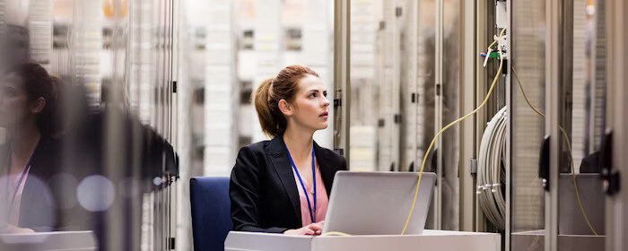 A woman working on a laptop in a server room, monitoring network connections.